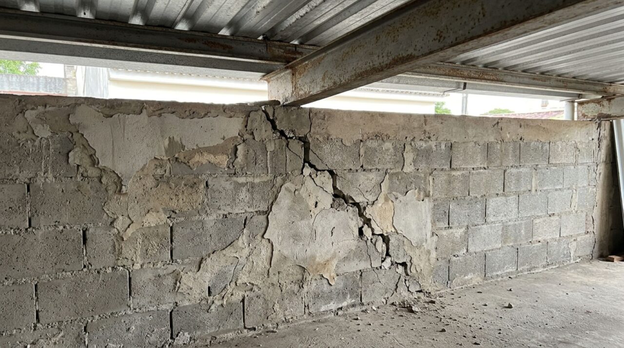 Muro de blocos de concreto com rachadura diagonal próxima a uma viga metálica que sustenta a cobertura de uma garagem.