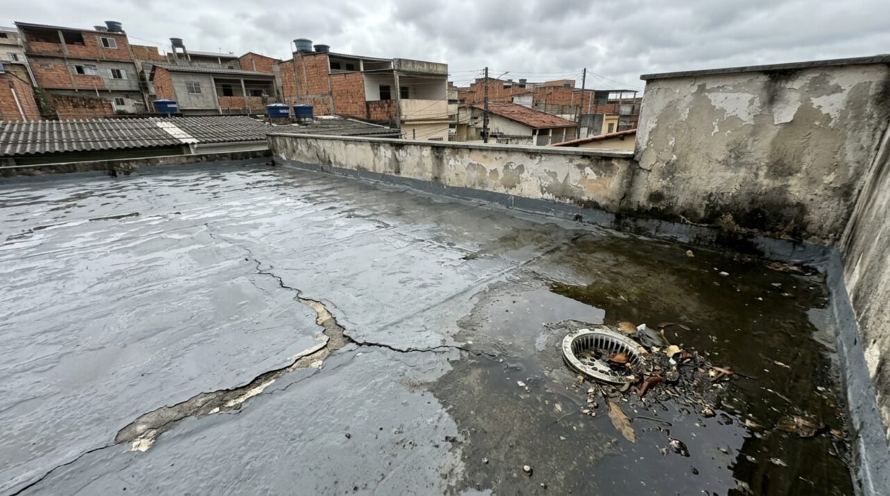 Laje impermeabilizada com trinca na superfície, poça de água da chuva e ralo parcialmente obstruído, mostrando possíveis causas de infiltração.