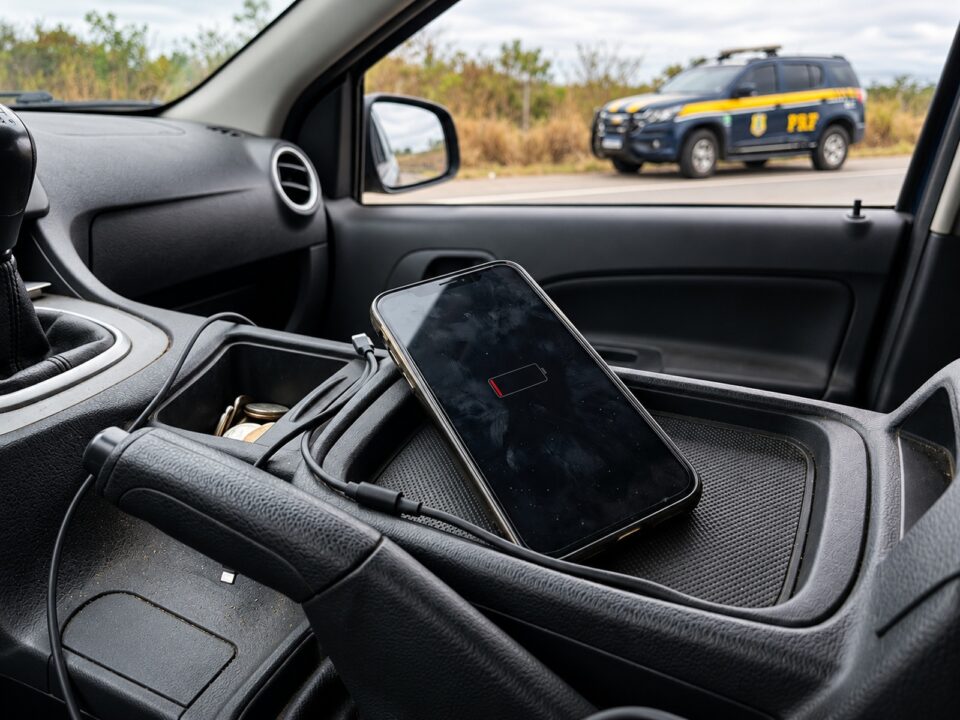 Celular com bateria descarregada no interior de um carro estacionado, com viatura da Polícia Rodoviária Federal ao fundo.